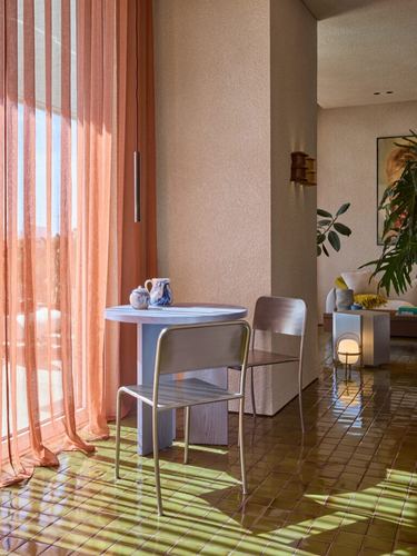 Small round table and metal chairs on terracotta tile floor beside sheer coral curtains