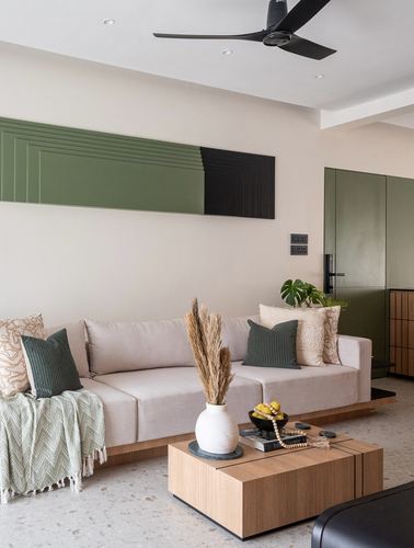 Beige sofa with green cushions faces a wooden coffee table beneath a green-black geometric wall panel and black ceiling fan