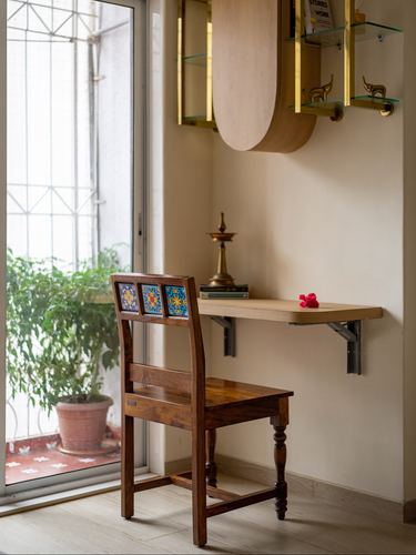 Wall-mounted wooden desk with brass lamp beside a carved wooden chair with painted tile inlay, near a sunlit balcony door