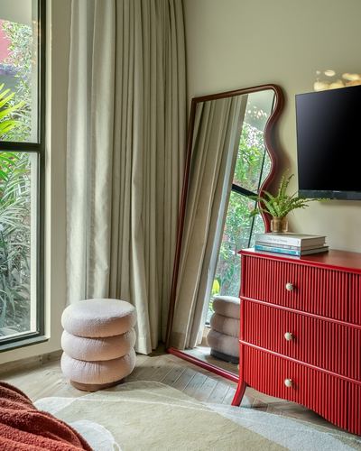 Red fluted dresser and wavy mirror beside curtained window with boucle pouf