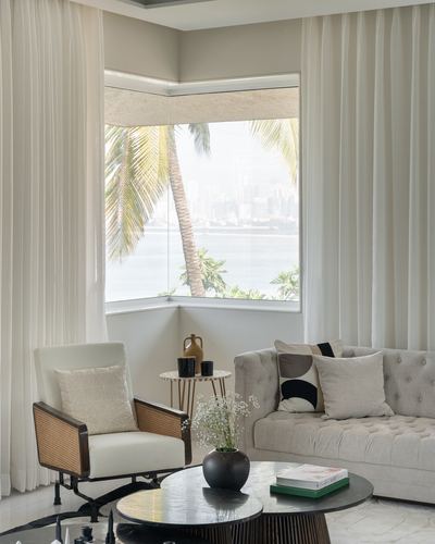Living room corner with sheer curtains, tufted sofa and cane chair framing a sea view window