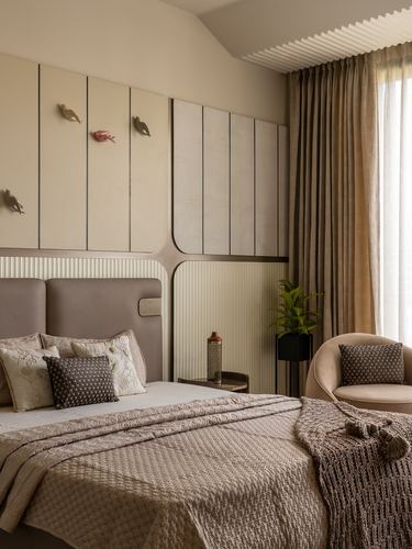 Bedroom with paneled wood wall, taupe upholstered headboard, curved chair and sheer-curtained window