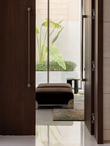 Dark wood pivot door opens onto a marble hallway with velvet ottoman and courtyard plant beyond