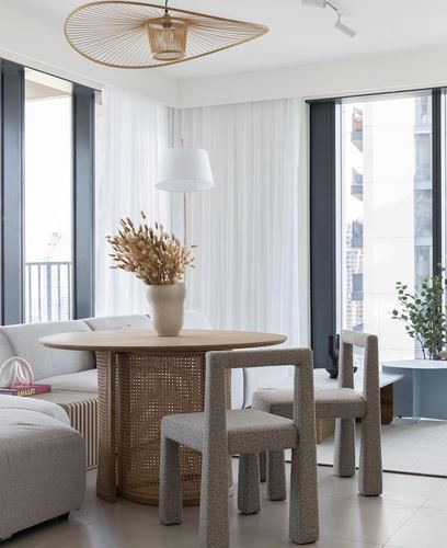 Round cane-base dining table with boucle chairs beside floor-to-ceiling windows in a bright Dubai apartment