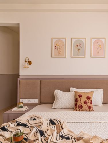 Bedroom with taupe upholstered headboard, three arched floral prints, brass sconce, and patterned throw on bed