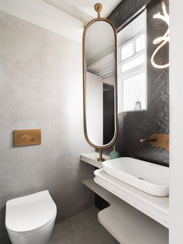 Small bathroom with grey stone walls, black herringbone tile, brass oval mirror and vessel sink