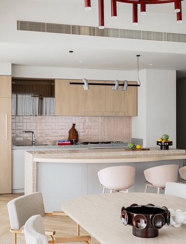 Open kitchen with pink brick-pattern tile backsplash, wood cabinetry, and a curved tiled island, seen past a dining table