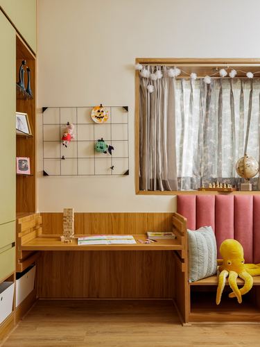 Child's study desk beside pink upholstered window seat with toys and curtains