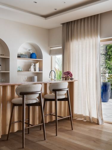 Kitchen counter with two wooden stools, arched shelving niches, and sheer curtains beside garden view
