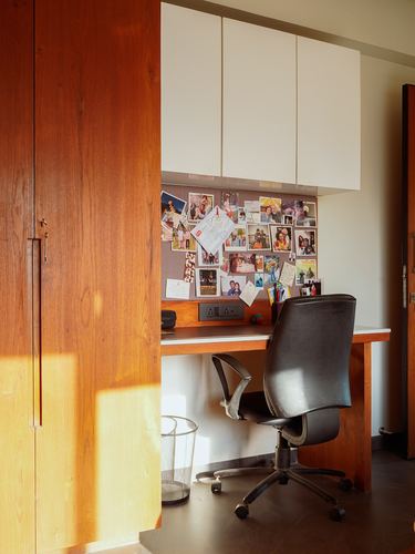 Sunlit study nook with wooden cabinet, white overhead storage, photo pinboard, desk and black office chair