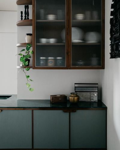Dark green stone counter beneath wooden cabinets with fluted glass doors and open shelving in a Chennai kitchen