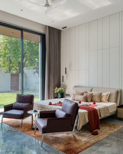 Bedroom with paneled wall, upholstered bed, and two burgundy leather armchairs facing a garden view