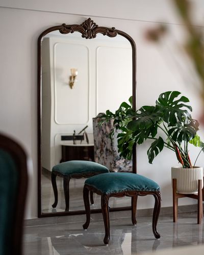 Antique arched mirror with carved crest beside teal velvet stools and a monstera plant on marble flooring.