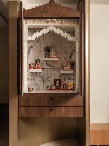 Wall-mounted wooden pooja unit with carved white arch, idols, and peacock-print wallpaper backdrop