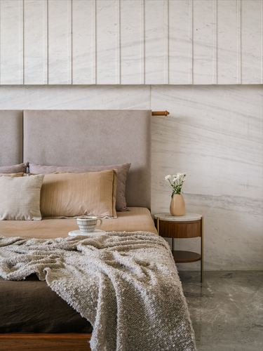 Bedroom corner with grey upholstered headboard, marble wall, wooden nightstand and knit throw