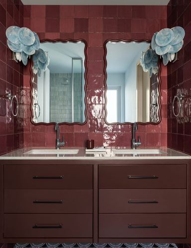Dark red tiled bathroom with double wavy mirrors, blue petal sconces, and maroon vanity