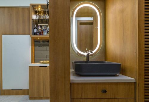 Wood-panelled vanity with backlit oval mirror and dark vessel sink beside a built-in bar nook