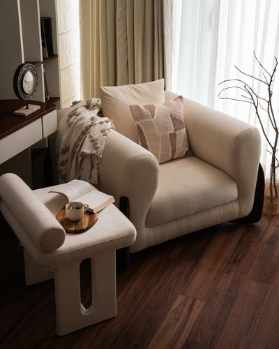 Cream armchair with throw and patterned cushion beside a cutout stool holding a book and coffee, dark wood floor.