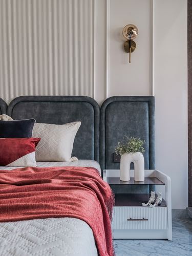 Bed with grey velvet arched headboard, red throw, and white bedside table with arched vase and wall sconce