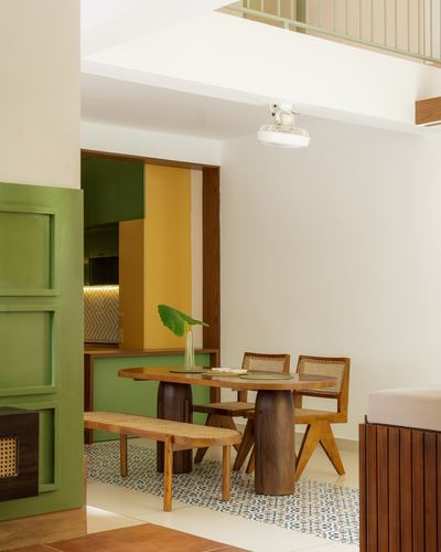 Dining nook with sculptural wood table, cane chairs and bench, opening to a green and yellow kitchen.
