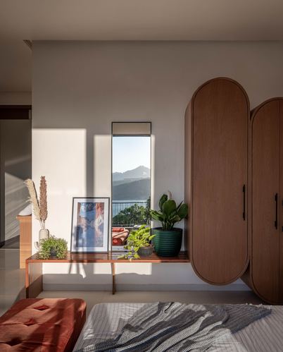 Sunlit bedroom corner with rounded wooden wardrobe, narrow window framing a lake view, and console with plants