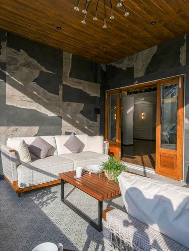 Covered patio with wicker sofas, wooden table, and stone-clad wall opening to an interior room.