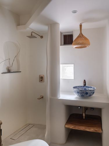 Curved plaster bathroom with brass shower fixtures and blue-and-white ceramic vessel sink under woven pendant