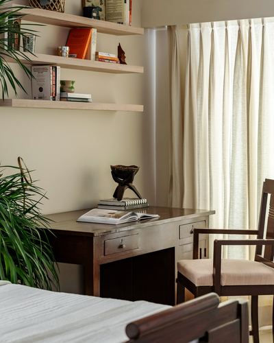 Wooden desk with books beneath floating shelves near a curtained window and cane armchair