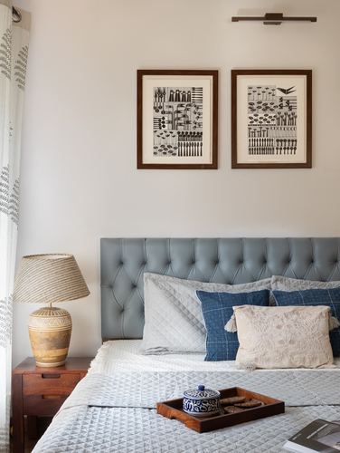 Bed with grey tufted headboard, layered blue and cream pillows, framed botanical prints, and wooden nightstand with ceramic lamp