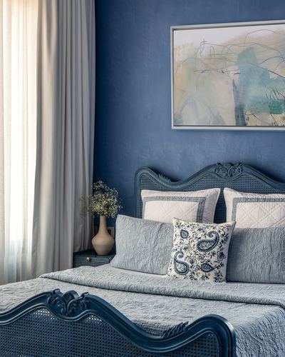 Blue bedroom with carved cane headboard, layered pillows, and abstract art on textured blue wall