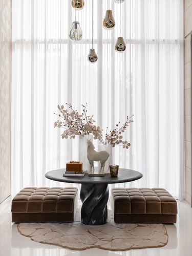 Foyer vignette with sculptural table, twisted base, flanked by brown velvet ottomans against sheer curtains