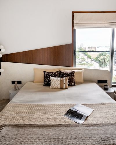 Bed with beige linens and cushions against a wood and upholstered headboard, city view through window
