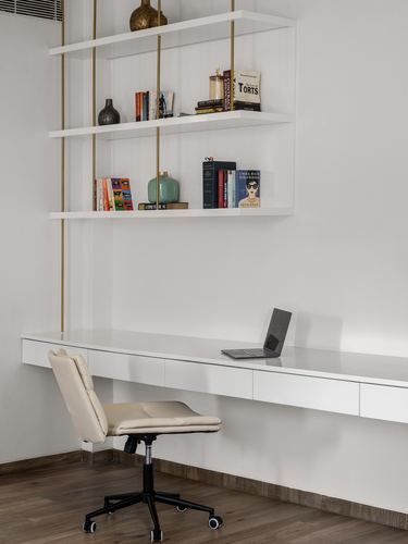 White floating desk and suspended brass-rod bookshelf in minimalist home study with cream chair