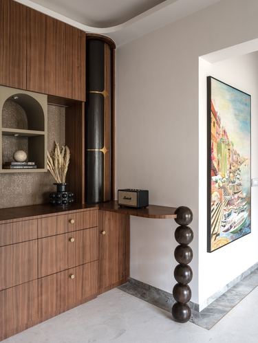 Wood cabinetry beside a black-gold cylindrical column and a sculptural chain-leg console table under a colorful framed artwork.
