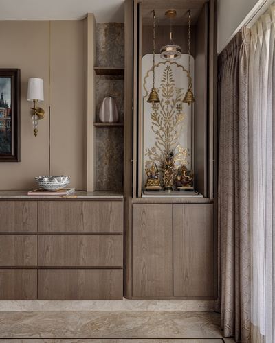 Wooden pooja unit niche with hanging brass bells and deity idols beside dresser and curtain