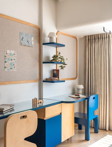 Child's study nook with blue plywood desk, cane-edged pinboards, and wall-mounted shelves against beige curtain