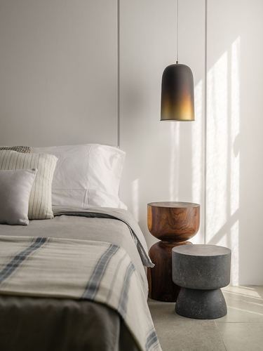 Bedroom corner with layered linen bedding, wood and cement hourglass side tables, and a dark pendant light in raking sunlight