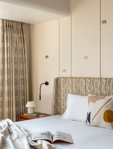 Bedroom with panelled cream wall, elephant motifs, patterned headboard, floral curtains and wooden nightstand