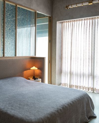 Minimalist bedroom with grey upholstered headboard, sheer curtains, and textured blue glass partition above bed