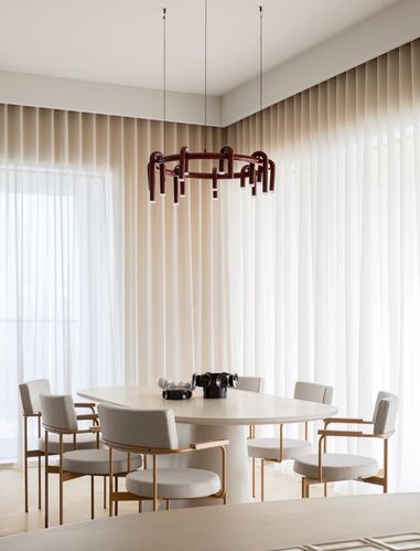Oval dining table with cream chairs beneath a burgundy sculptural pendant, sheer curtains behind