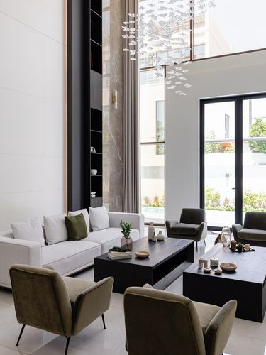 Double-height living room with olive armchairs, black coffee tables, and a cascading chandelier by full-height windows