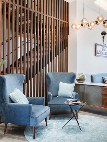 Two blue wingback chairs before a wood-slatted staircase screen and glass pendant cluster light