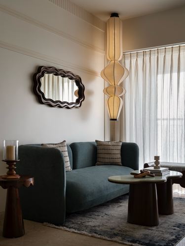 Green velvet sofa beneath wavy mirror with sculptural pendant lamp beside sheer curtains