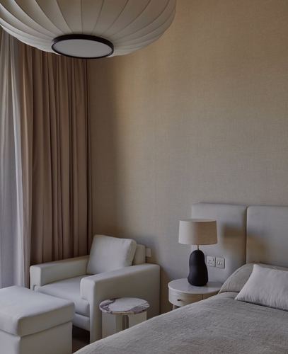 Beige minimalist bedroom corner with armchair, ottoman, round marble side table, and upholstered bed