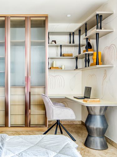 Corner study nook with floating shelves, patterned wall, desk, velvet chair and metal stool beside a mirrored wardrobe