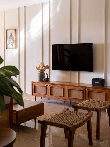 Wooden TV console with cane doors below wall-mounted TV, woven stools, fluted wall panels, sunlight streaks