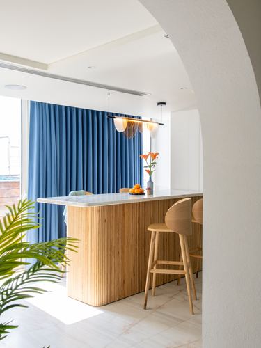 Fluted wood kitchen island with marble top under pendant lights, blue curtain backdrop, seen through arched wall.