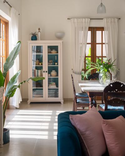 Sunlit living-dining room with white curtains, teal sofa, and glass display cabinet in Goa villa