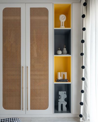 Cane-front wardrobe with brass handles beside mustard and grey shelving niches holding sculptural objects and books.