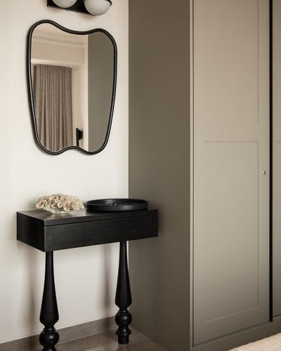 Black turned-leg console with organic mirror against cream wall beside olive wardrobe
